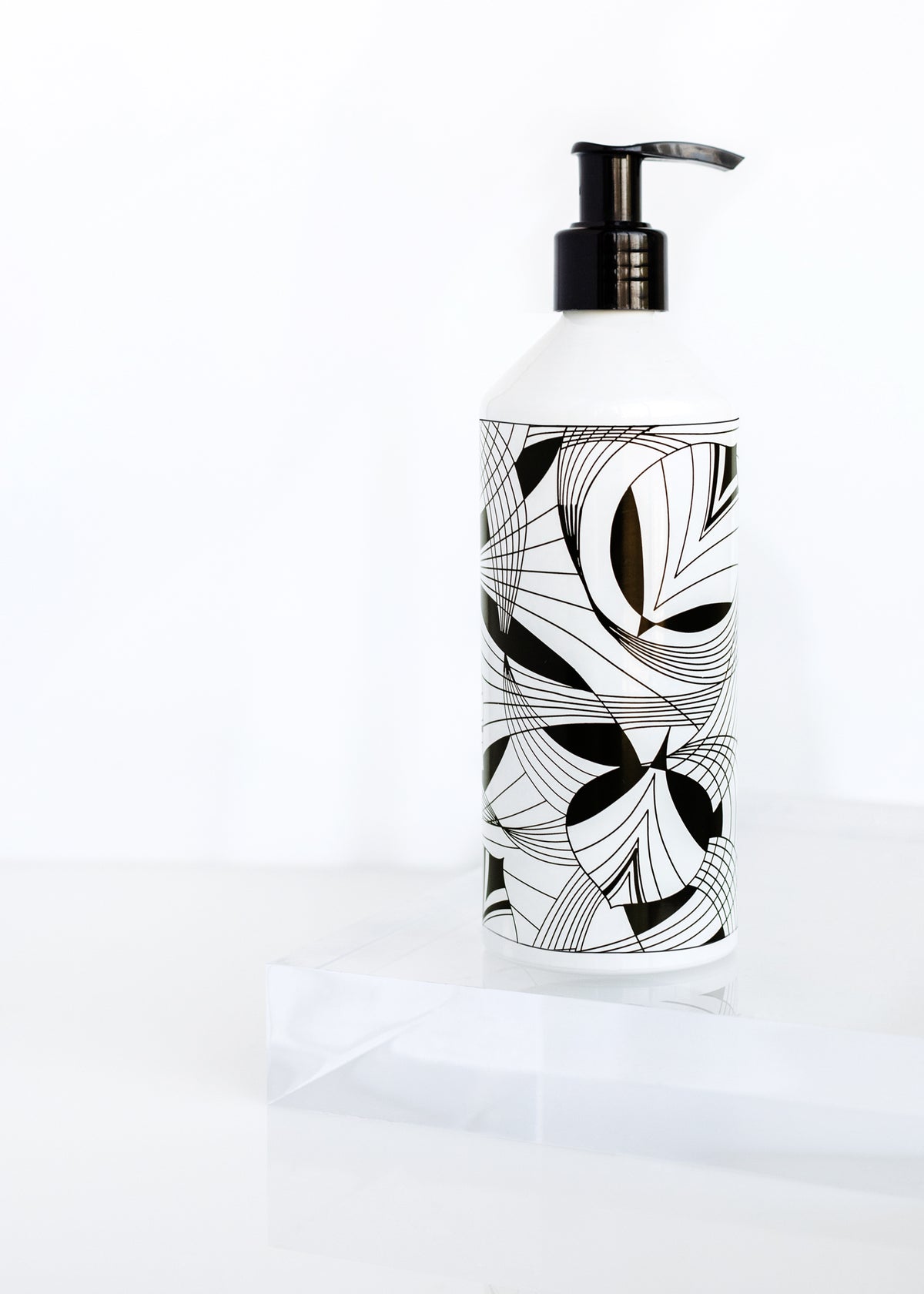 A Margot Elena lotion dispenser with a modern black and white geometric design, featuring vegan skincare ingredients, placed on a clear surface against a white background.