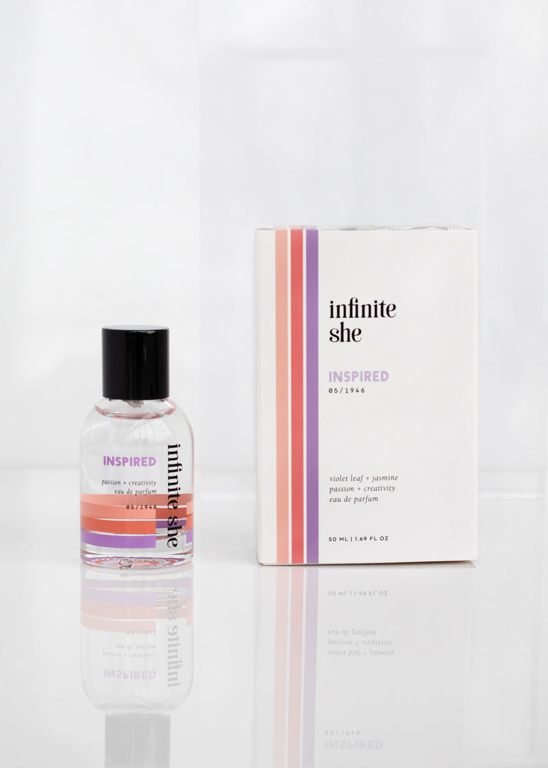A bottle and a box of Margot Elena's "Infinite She Inspired Eau de Parfum" vegan fragrance on a reflective surface, featuring pink liquid, with a description of scent notes including jasmine blossoms and cedarwood.