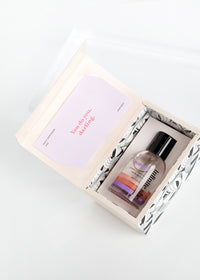 An open gift box containing a bottle of Margot Elena's Infinite She Inspired Eau de Parfum and a makeup palette, with a visible note saying "you do you, beautiful!" on a clean white background.