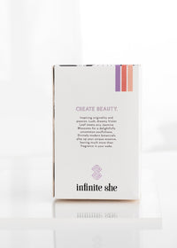 A white product box for Margot Elena's "Infinite She Inspired Eau de Parfum" vegan fragrance, featuring elegant text and colorful stripes on a bright, reflective surface.