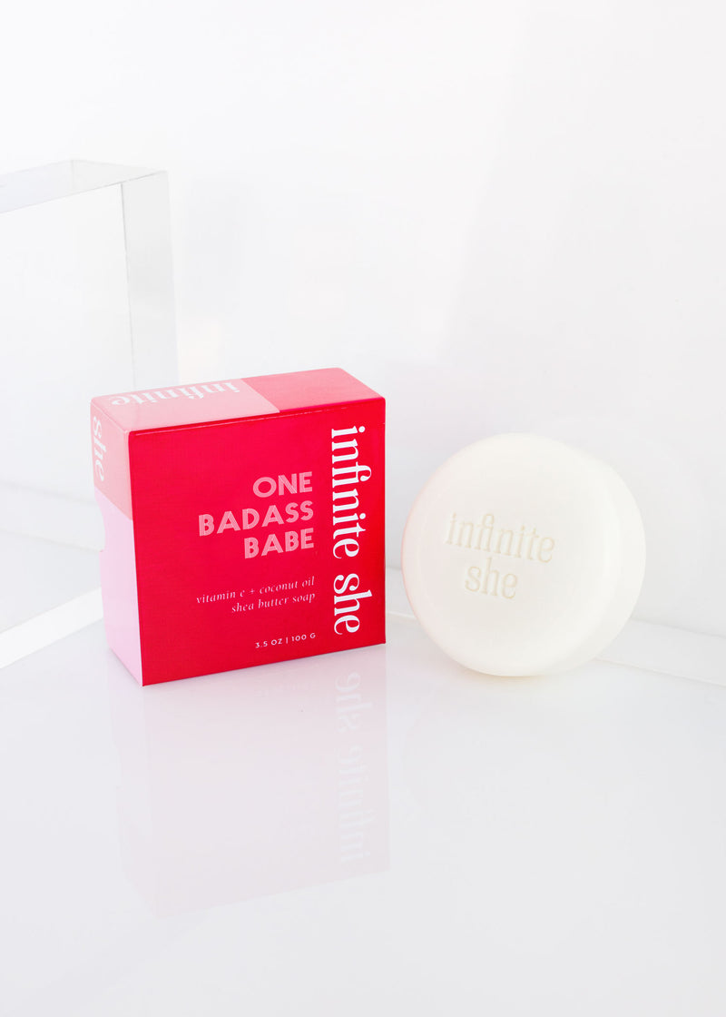 A bright pink box with the text "one badass babe" next to a white round container labeled "infinite she" against a clean, white background, featuring Margot Elena's Infinite She One Badass Babe Shea Butter Soap.