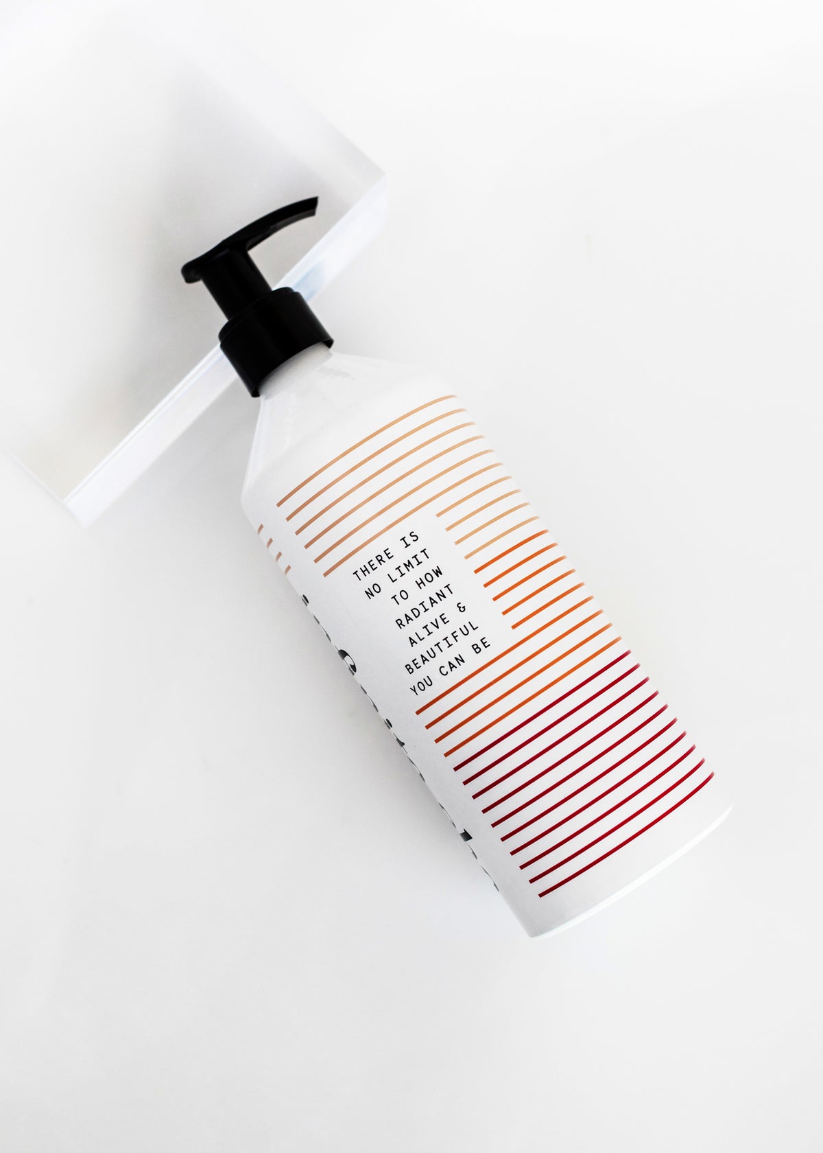 A white Infinite She Vibrant Hydrating Body Lotion bottle with a black pump, lying on a white surface. The bottle features orange diagonal stripes and text that reads, "there is no limit to how radiant, alive, and irresistible you" by Margot Elena.