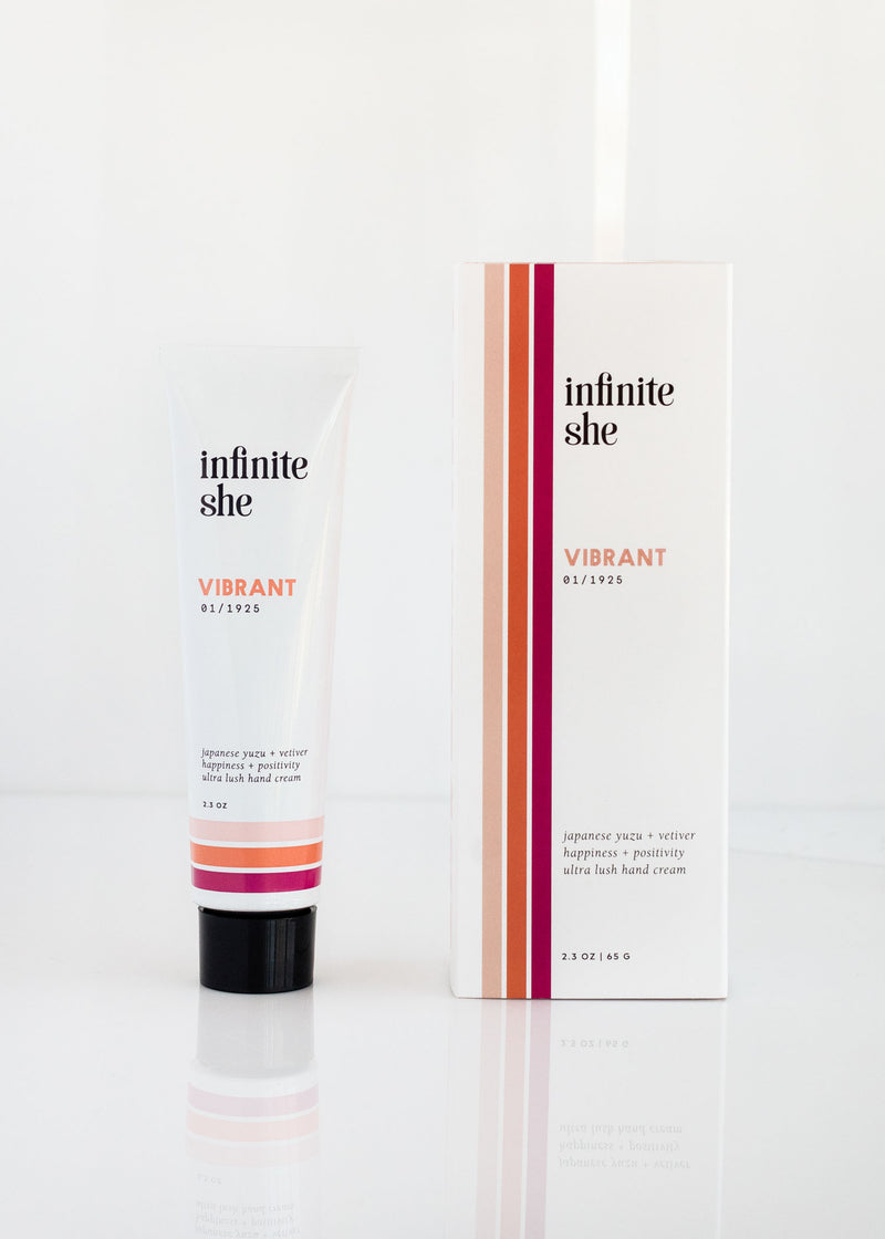A tube of Margot Elena's 'Infinite She Vibrant Ultra Lush Hand Cream' beside its packaging box, both featuring clean, modern design with red and black accents on a white background. The product is enhanced with Shea + Mango But