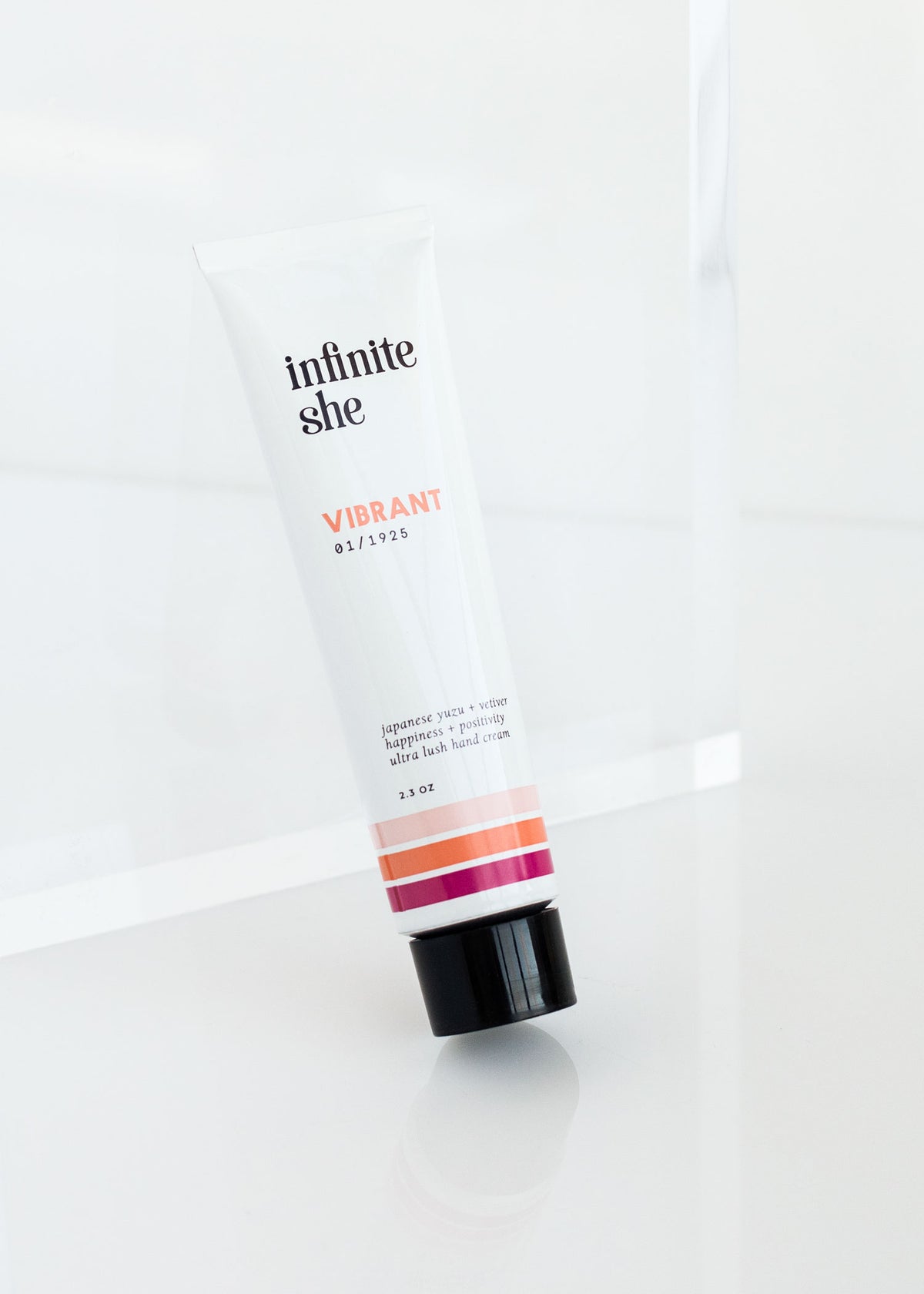 A tube of Margot Elena's "Infinite She Vibrant Ultra Lush Hand Cream" placed upright on a shiny, reflective white surface, with a minimalist background. The tube has a white body with black cap and pink-striped design.
