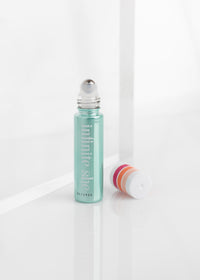 A clear roll-on bottle of Margot Elena's Infinite She Vibrant Rollerball Eau de Parfum with blue-green liquid next to its cap on a reflective white surface. The label on the bottle reads "Infinite She," featuring Japanese Yuzu.