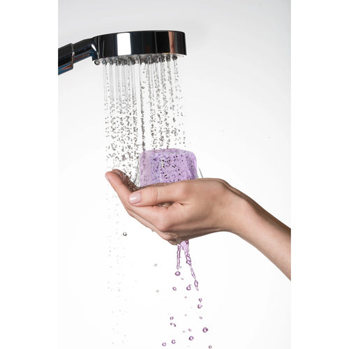 A hand holds a Lizush Lavender Shower Steamer under a running shower, with water streaming down and droplets splashing around.