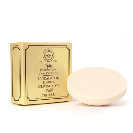 A round bar of Taylor of Old Bond Street Sandalwood Shave Bowl Refill beside its gold-colored box labeled "Taylor of Old Bond Street, Sandalwood Herbal Shaving Soap Refill, 100g 3