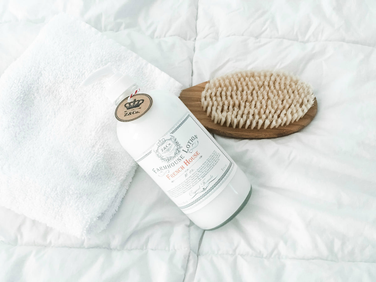 A bottle of Z&Co. French House Farmhouse Lotion, with natural ingredients, lies beside a wooden body brush on a soft, wrinkled white bedsheet. A fluffy white towel is partially visible. The setting suggests a calm, soothing atmosphere.