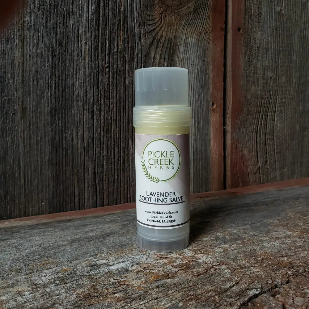 A stick of Pickle Creek Herbs Lavender Soothing Salve - 2.25 oz resting upright against a rustic wooden background, featuring clear labels with the product name and ingredients.