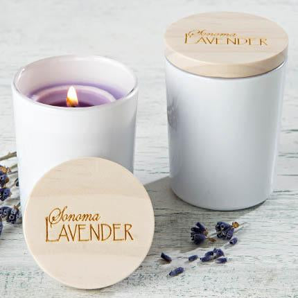 A Sonoma Lavender soy wax candle with a lit wick in a white ceramic container, next to a closed candle tin labeled "Sonoma Lavender," surrounded by scattered lavender buds on a wooden surface.