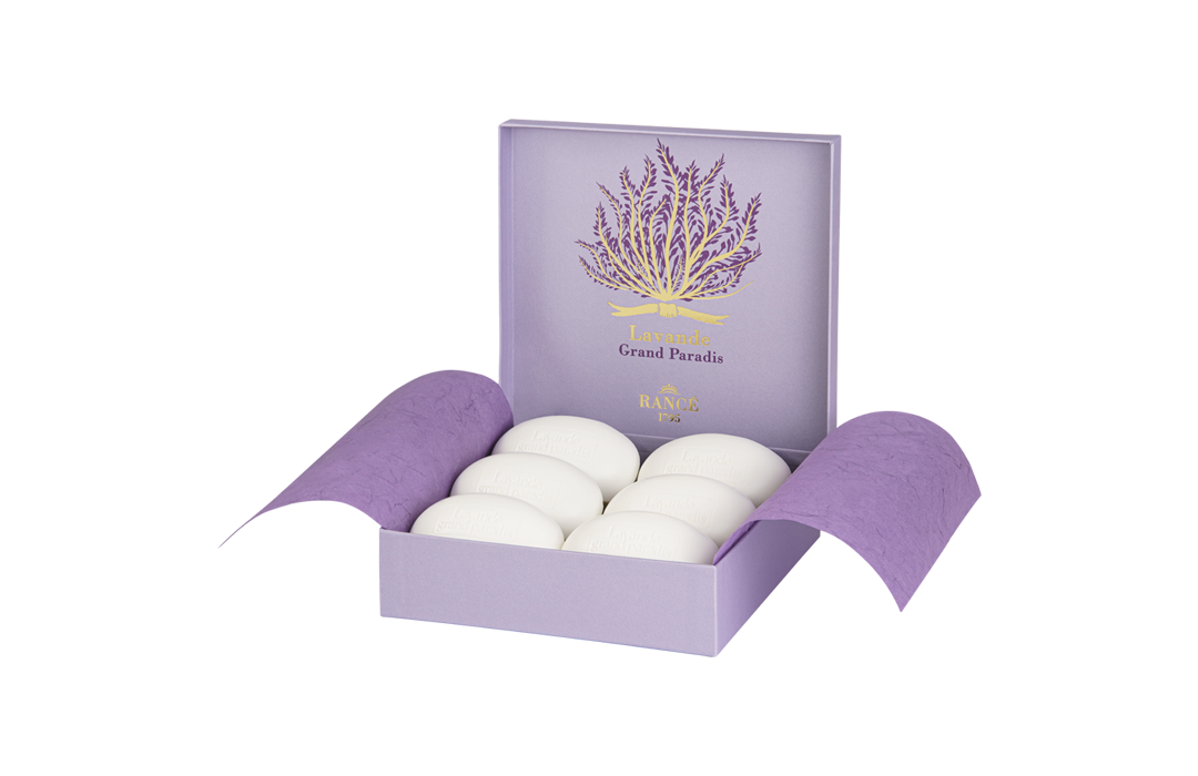A purple and white box with six white eggs displayed, accompanied by a purple book titled "Rancé Classic Soap - Lavande Grand Paradis." The background features soft lavender stripes and a hint of Rancé tradition.