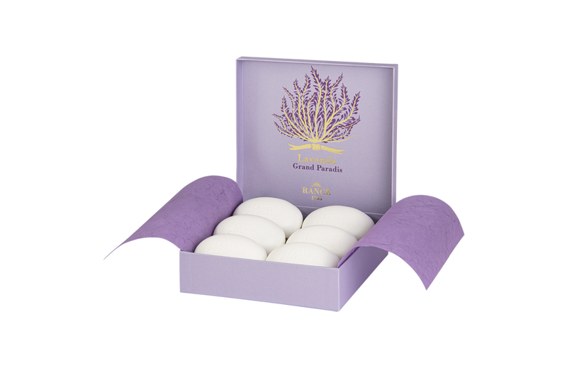 A purple and white box with six white eggs displayed, accompanied by a purple book titled "Rancé Classic Soap - Lavande Grand Paradis." The background features soft lavender stripes and a hint of Rancé tradition.