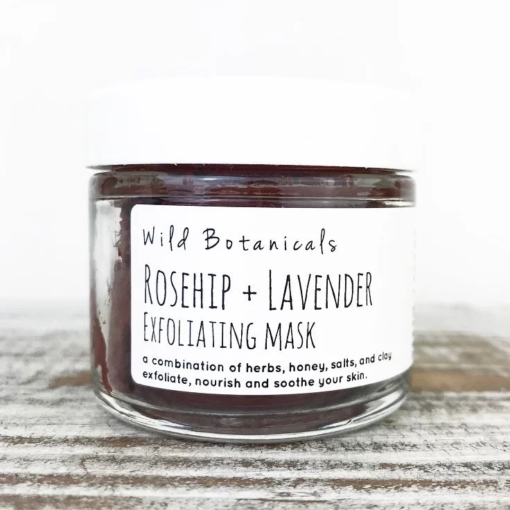 A glass jar of Wild Botanicals Rosehip and Lavender Exfoliating Mask on a light wooden surface, with a label describing its natural ingredients including Aloe Vera powder.