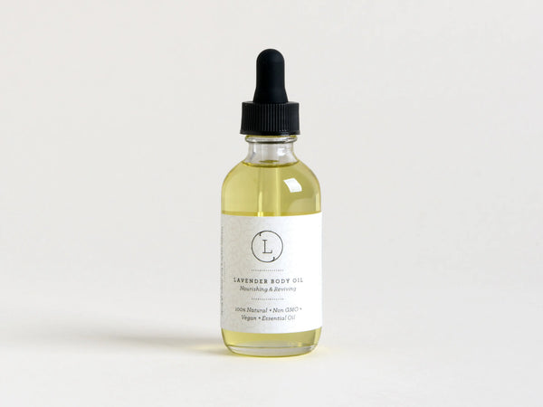A clear glass bottle containing Lizush Lavender Body Oil with a dropper, labeled with white text stating "moisturizing lavender body oil, nourishing & reviving." The background is plain and light-colored.
