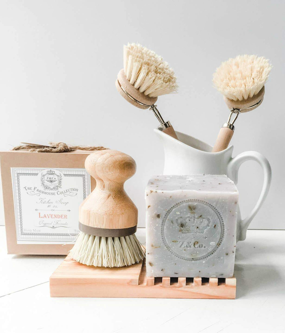 A neat display of eco-friendly cleaning supplies including two wooden scrub brushes, a bar of Z&Co. Lavender Farmhouse Solid-Block Dish Soap, and a soap dish, arranged next to a white ceramic pitcher on a light background.