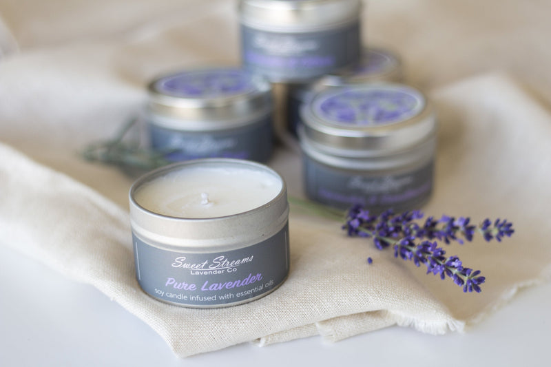An arrangement of Sweet Streams Lavender Co. - Pure Lavender Soy Travel Tin Candles in silver travel tins with labels, displayed on a beige cloth alongside some fresh lavender sprigs. The foreground focuses on one open candle.