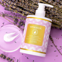 A bottle of Spongellé French Lavender Body Lotion with Shea Butter on a purple background, accompanied by lavender stalks and a dab of cream on a transparent dish.