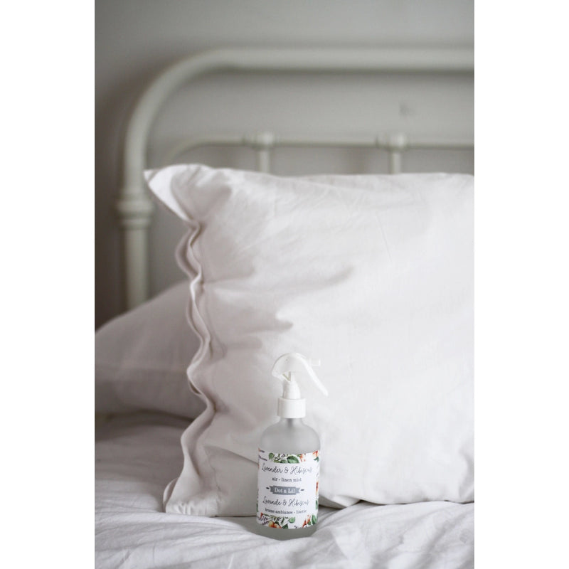 A white pillow on a bed with a decorative Dot & Lil Lavender + Hibiscus Linen + Air Mist spray bottle placed in front of it, set against a soft, light background.
