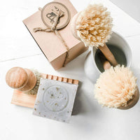 Top-down view of eco-friendly bathroom accessories including wooden brushes, a Z&Co. Lavender Farmhouse Solid-Block Dish Soap, and a small towel, all arranged on a white surface.