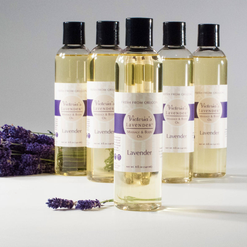 Five bottles of Victoria's Lavender Lavender Massage & Body Oil, labeled and displayed in a row on a light background, with fresh lavender sprigs in the foreground.