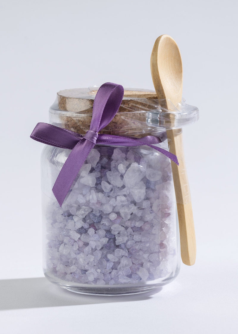 A Sonoma Lavender Bath Salts in Honey Jar filled with purple and white sea bath salts, sealed with a cork lid and decorated with a purple ribbon. A wooden spoon is attached to the side of the jar.