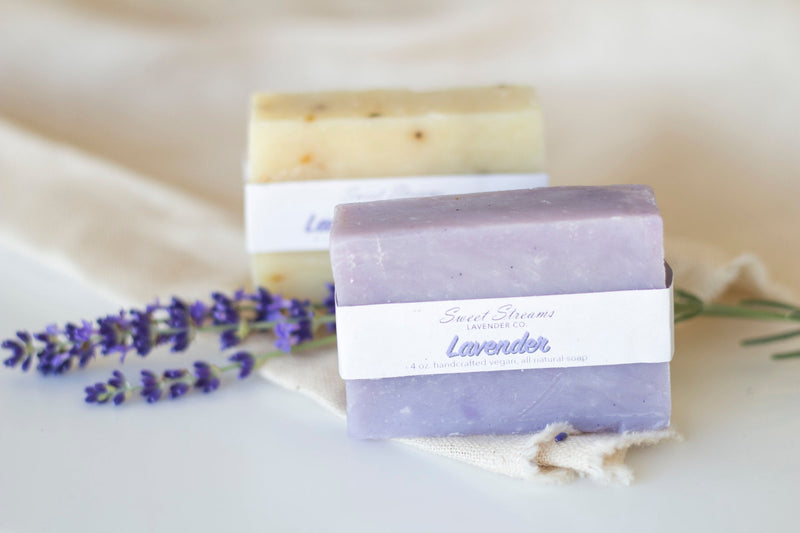 Two bars of lavender soap labeled "Sweet Streams Lavender Co. - Lavender Soap," with fresh lavender sprigs, on a light fabric background. The upper bar has visible petals mixed in.