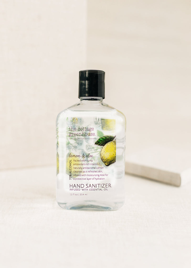 A clear bottle of Margot Elena's "The Cottage Greenhouse Lemon & Aloe Hand Sanitizer Gel" sits on a textured white surface, with a soft background. The label shows images of lemon slices and highlights its antibacterial Lemon properties.