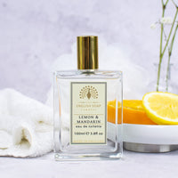 A bottle of The English Soap Co. Vintage Lemon & Mandarin EDT displayed with sliced lemon, white flowers, and a fluffy white towel on a light background.