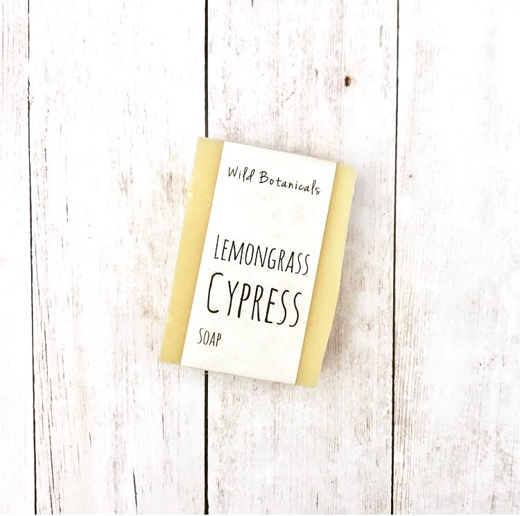 A bar of Wild Botanicals Lemongrass Cypress Soap, featuring essential oils, rests on a white wooden surface, displaying a minimalist and natural design.