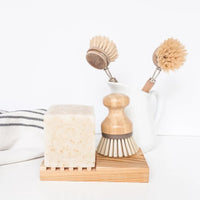 Two Saucepan Brushes in a white cup next to a bar of Z&Co. Lemon Farmhouse Solid-Block Dish Soap on a wooden stand, all against a clean, white background. A striped cloth is partially visible.