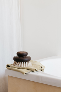A Andrée Jardin Heritage Ash Wood Large Body Brush resting on a pale fabric next to a white bathtub, against a softly lit cream-colored background.