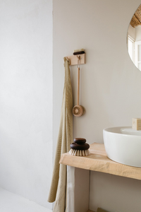 A minimalist bathroom corner featuring a wooden shelf with a white basin, and Andrée Jardin Heritage Ash Wood Large Body Brushes hung on the wall beside a folded beige towel.