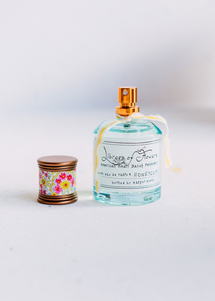 A perfume bottle labeled "Library of Flowers Honeycomb Eau de Parfum" next to a small floral-patterned lid against a white background. The bottle features elegant script and a visible spray nozzle by Margot Elena.