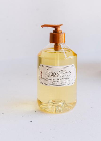 Clear glass bottle with honey-colored Library of Flowers Honeycomb Shower Gel featuring a pump dispenser and labeled "Margot Elena, library of flowers american small batch perfumery, honeycomb with Shea Butter." Set against a plain white background.