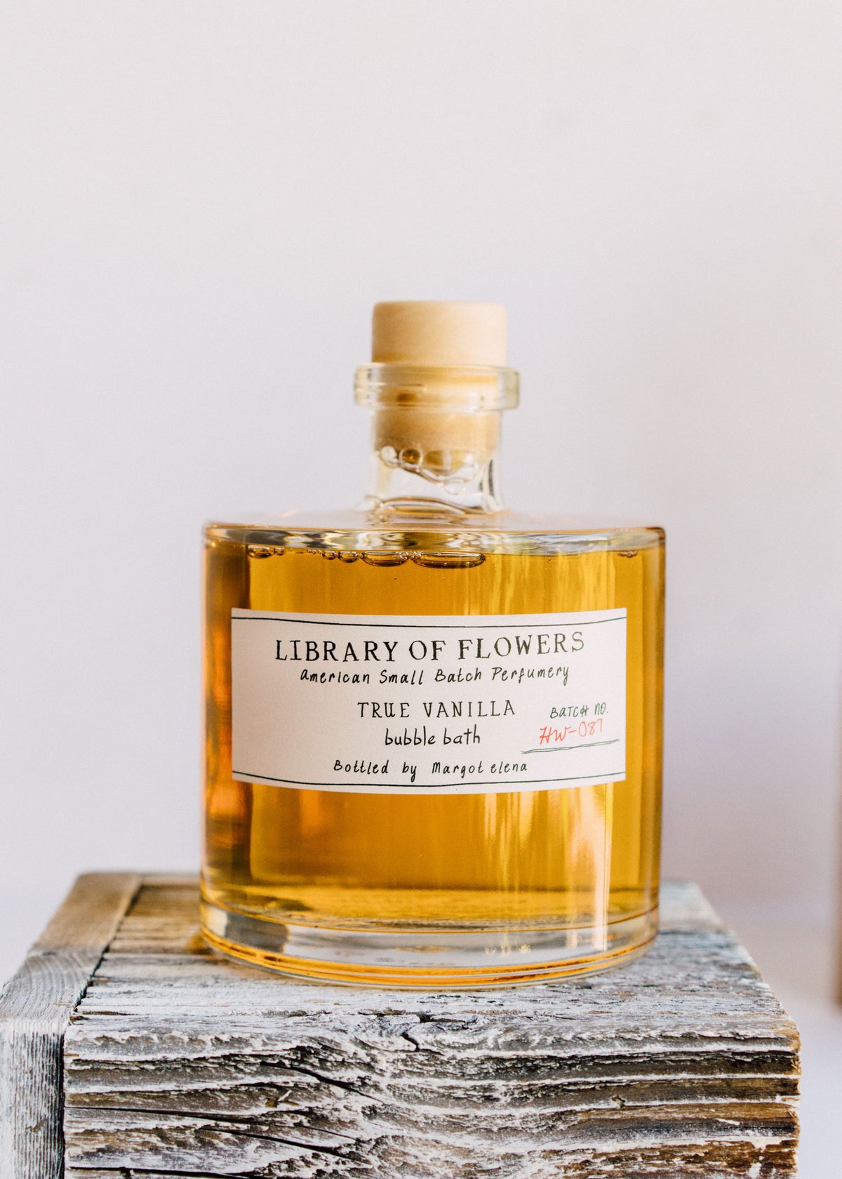 A clear glass bottle of Margot Elena's Library of Flowers True Vanilla Bubble Bath atop a rustic wooden block, positioning the well-lit label front and center against a neutral background.