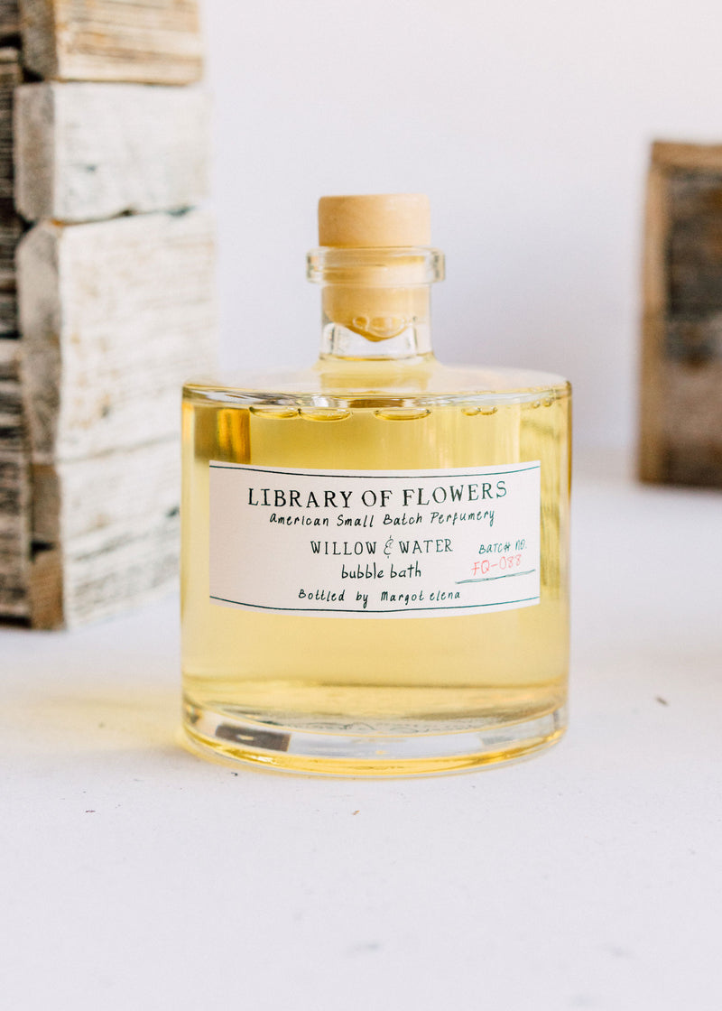 A glass bottle of Margot Elena brand Library of Flowers Willow & Water Bubble Bath stands against a blurred background of pale stones. The bottle contains a yellowish liquid with hints of green tea.