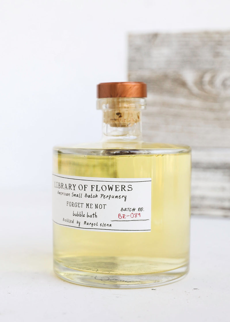 A glass bottle of Margot Elena's Library of Flowers Forget Me Not Bubble Bath with a cork lid, placed against a blurred white and wooden background, features silky suds.