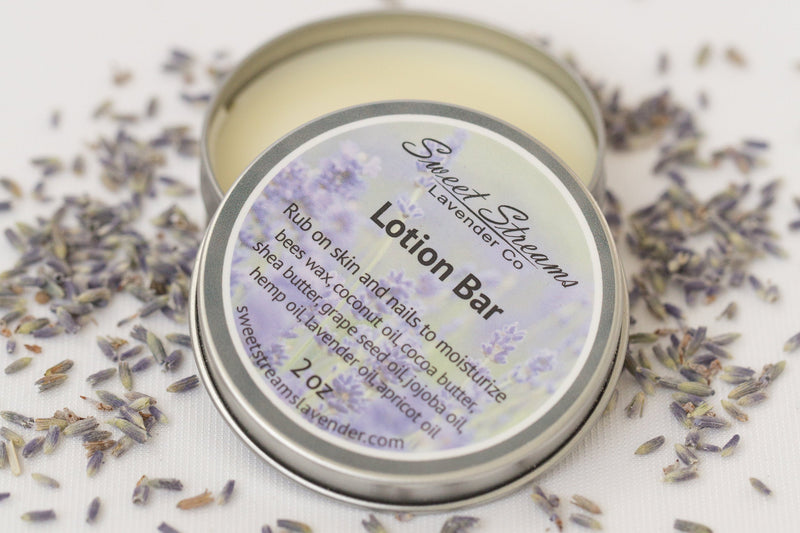 A close-up image of an open Lavender Lotion Bar in a metal tin, surrounded by scattered lavender flowers and enriched with cocoa butter, with a label that includes product details and ingredients. Brand: Sweet Streams Lavender Co.