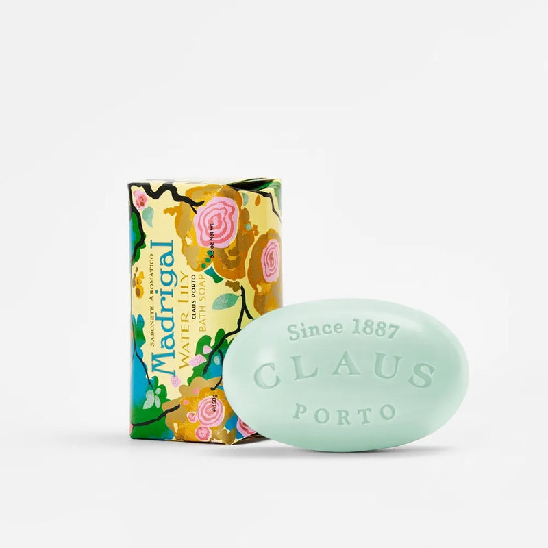 A Claus Porto 1887 Madrigal Water Lily soap bar next to its colorful floral packaging. The light green soap enriched with shea oil is engraved with "Claus Porto" and rests against a white background.