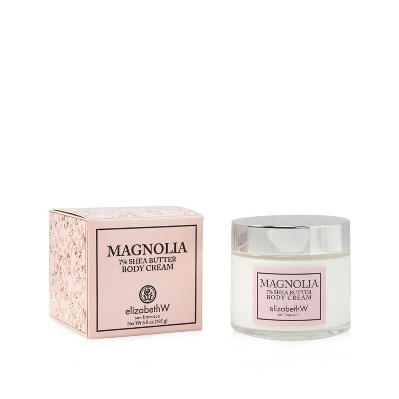 A jar of elizabeth W Signature Magnolia Body Cream hydrating cream next to its pink packaging box on a white background. The jar is clear with a white label and cream visible inside.