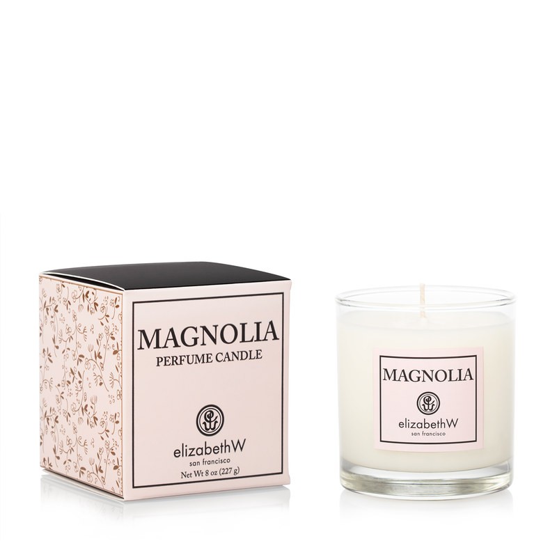 A white elizabeth W Signature Magnolia Candle, presented next to its elegant packaging with floral design. The candle is in a simple, clear glass container with the logo and fragrance name.
