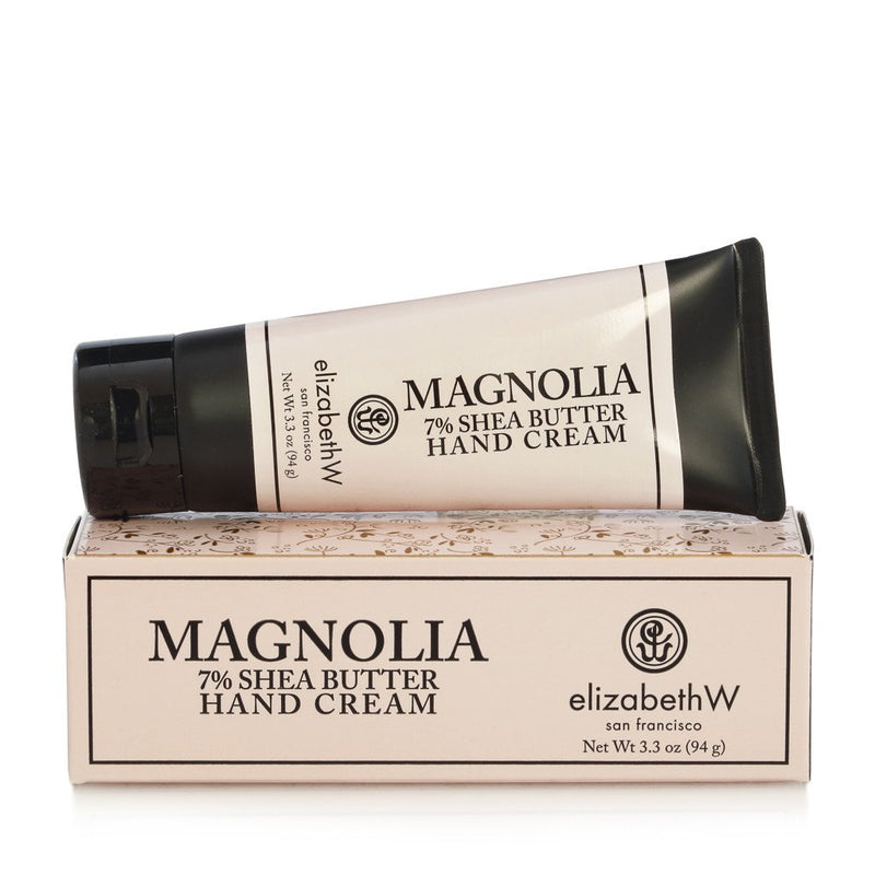 A tube of Elizabeth W Signature Magnolia Hand Cream with 7% shea butter, intensely hydrating, placed on top of its packaging box, isolated on a white background.