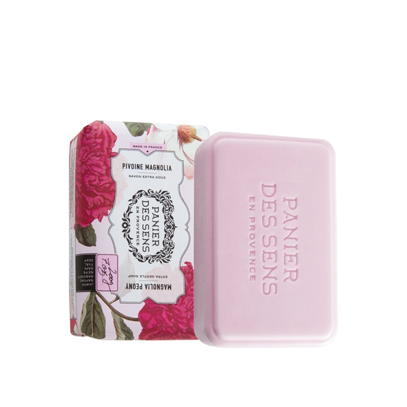 A pink bar of Panier Des Sens Extra-Soft Vegetable Soap labeled "panier des sens" next to its floral-patterned box with "Magnolia Peony" written on it, on a white background.
