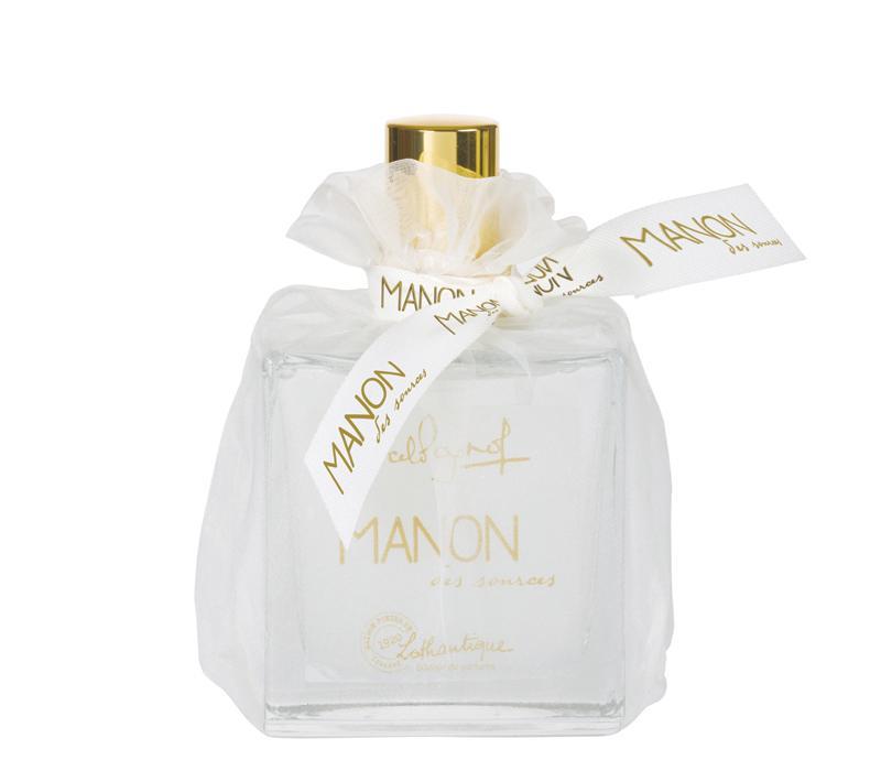 A clear glass Lothantique perfume bottle with a gold cap, adorned with a white bow and a small label featuring elegant gold script writing, encasing Lothantique Manon des Sources Eau de Toilette.