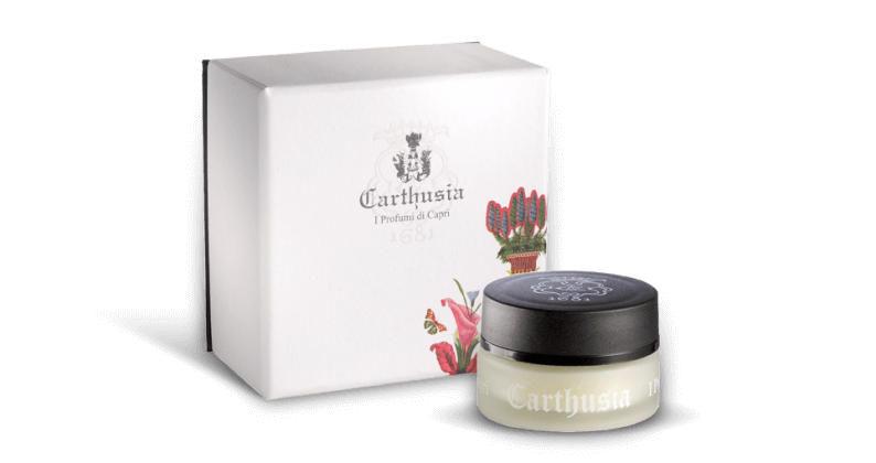 A small, open jar of Carthusia I Profumi de Capri solid perfume with Mediterraneo fragrance next to its white packaging box adorned with a floral design and the Carthusia logo. The setting is simple.