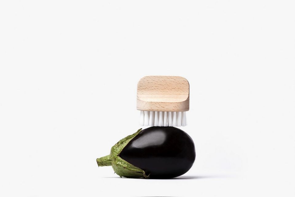A small Andrée Jardin "Canot" Vegetable Brush Medium with a natural wooden top and white bristles is perched atop a shiny, dark purple eggplant with a green stem, on a plain white background.