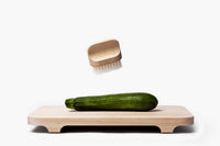 A zucchini resting on a wooden cutting board with a small Andrée Jardin "Canot" Vegetable Brush Medium with white bristles floating above it, both set against a white background.