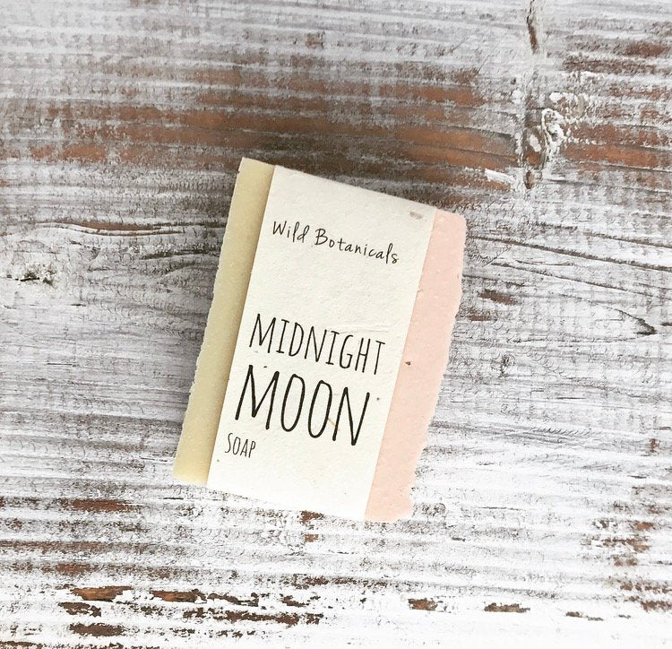 A bar of Wild Botanicals Midnight Moon Soap, handmade vegan soap from Wild Botanicals, displayed on a rustic wooden surface. The soap packaging features a neutral color palette.