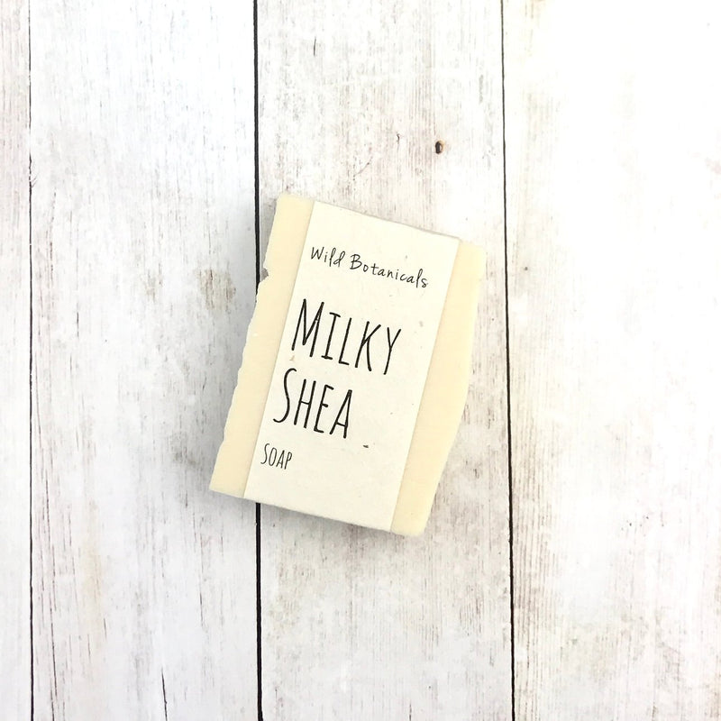 A bar of Wild Botanicals Milky Shea Soap with organic ingredients by Wild Botanicals lying on a white wooden surface.