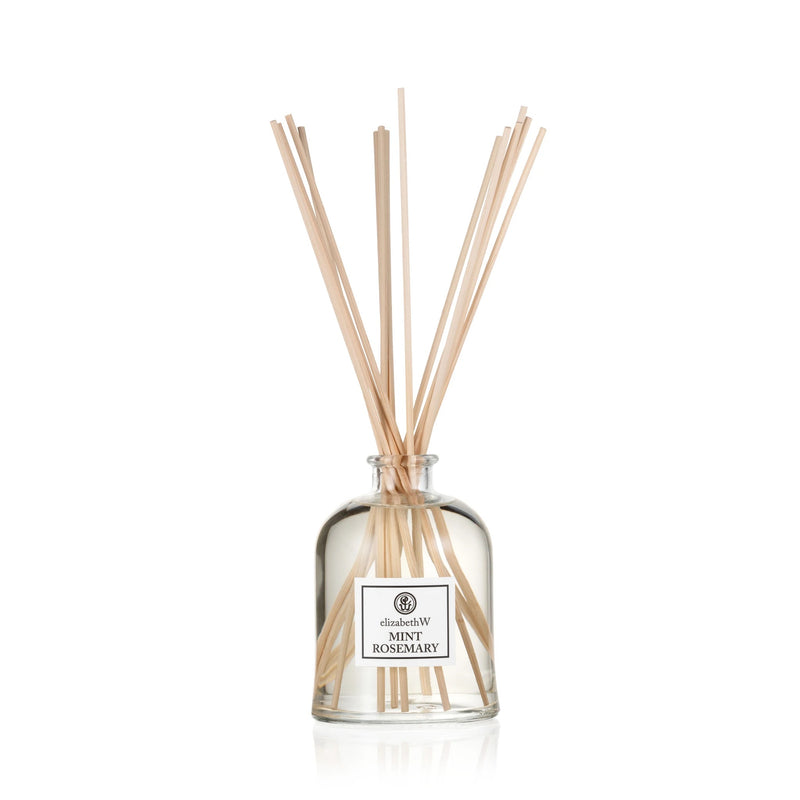 A clear glass fragrance diffuser bottle labeled "elizabeth W Purely Essential Mint-Rosemary Diffuser" with several light brown reed sticks inserted, standing against a white background.
